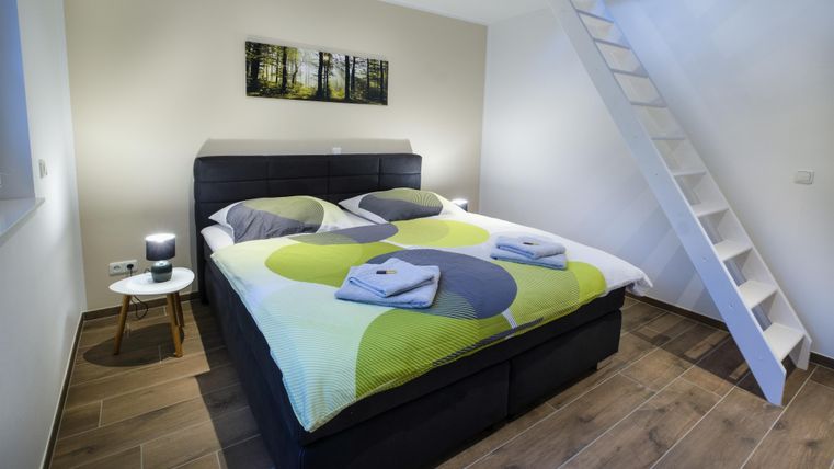 Ein modernes Schlafzimmer mit einem großen Bett, bunten Bettwäsche und einem kleinen Nachttisch. Eine Treppe führt zu einem höheren Bereich des Raumes.