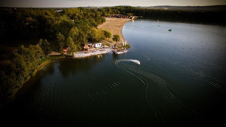 Ein ruhiger See mit einer kleinen Strandfläche und umgeben von Bäumen. In der Nähe ist ein Boot zu sehen, das über das Wasser gleitet.