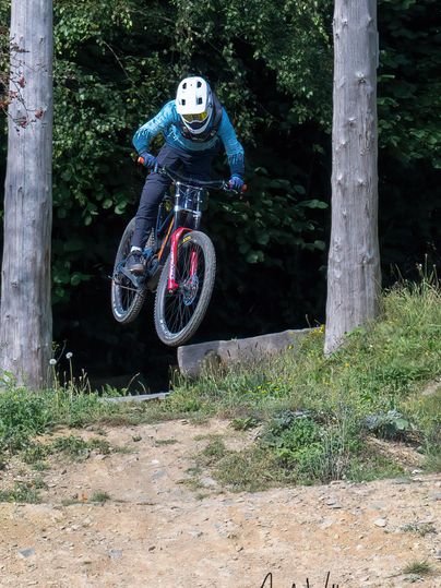Ein Fahrradfahrer springt über eine Rampe im Wald. Der Fahrer trägt einen Helm undenge Kleidung.