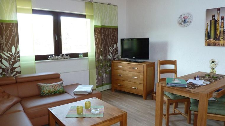 Ein gemütlicher Wohnbereich mit einem braunen Sofa, einem Tisch aus Holz und einer TV-Einheit. Helle Vorhänge und eine ruhige Farbpalette schaffen eine einladende Atmosphäre.