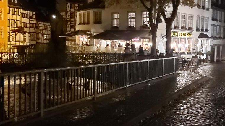 Eine ruhige Straße bei Nacht mit beleuchteten Gebäuden und einem kleinen Kanal. Am Rand steht ein Geländer und die Pflastersteine glänzen vom Regen.