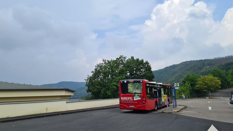 Ein roter Bus steht an einer Haltestelle. Im Hintergrund sind grüne Berge und ein bewölkter Himmel zu sehen.