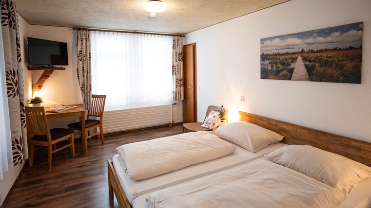 Ein gemütliches Schlafzimmer mit einem Doppelbett und einem kleinen Tisch. Die großen Fenster lassen viel Licht herein, und ein Fernseher ist an der Wand montiert.