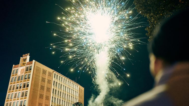 Ein Feuerwerk erleuchtet den Nachthimmel über einem Gebäude. Auf der gegenüberliegenden Seite ist eine Gruppe von Menschen zu sehen, die das Spektakel beobachten.