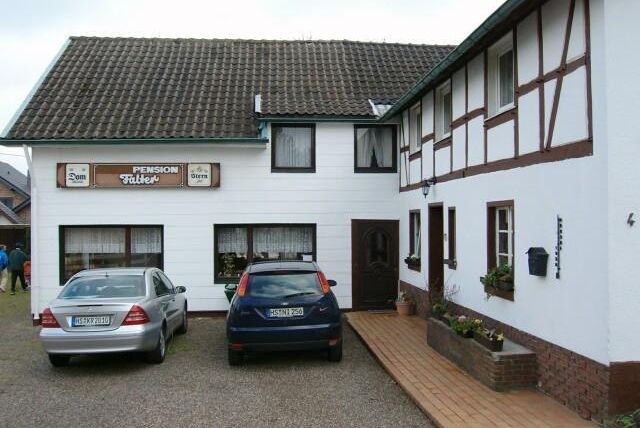 Ein Gasthaus mit einer Mischung aus modernem und traditionellem Baustil. Zwei Autos stehen vor dem Eingang auf einem gepflasterten Weg.