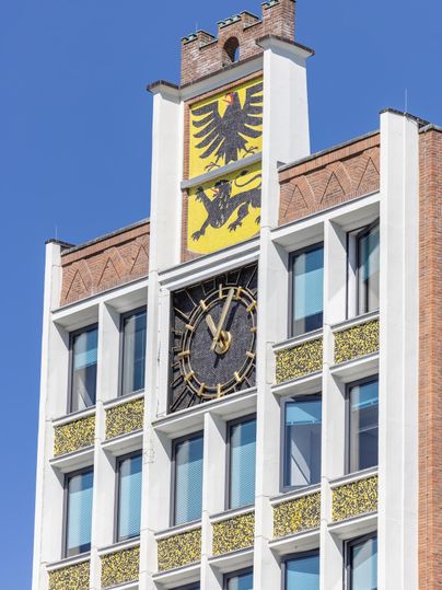 Ein historisches Gebäude mit einem großen Ziffernblatt und dem Wappen auf der Spitze. Der klare blaue Himmel sorgt für einen schönen Kontrast.