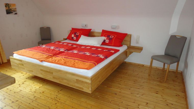 Ein modernes Schlafzimmer mit einem großen Holzbett und bunten Kissen. Der Boden ist aus Holz und ein Stuhl steht neben dem Bett.