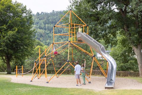 Ein farbenfrohes Klettergerüst mit einer Rutsche steht in einem Park. Im Hintergrund sind Bäume und sanfte Hügel zu sehen.