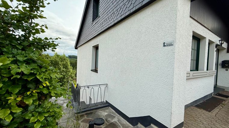 Ein modernes Haus mit weißer Fassade und grauem Dach. Im Vordergrund sind einige Pflanzen und ein gepflasterter Weg sichtbar.
