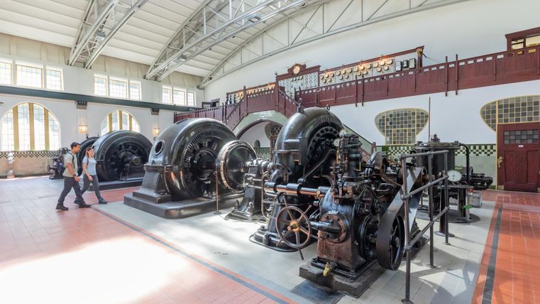 Ein historisches Kraftwerk mit großen Maschinen und einem hellen, offenen Raum. Zwei Personen gehen durch die Ausstellung.