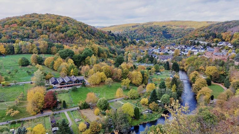 Eine malerische Landschaft mit bunten Herbstbäumen und einem ruhigen Fluss. Im Hintergrund ist ein kleines Dorf in den sanften Hügeln zu sehen.