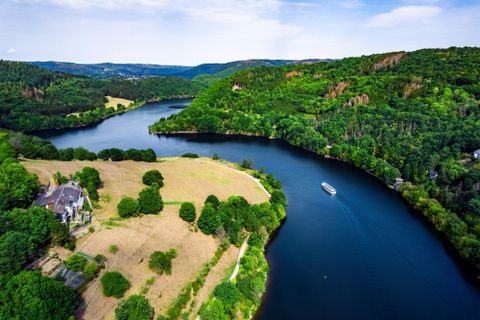Eine malerische Landschaft mit einem Fluss, umgeben von grünen Hügeln und Wäldern. Ein kleines Boot fährt ruhig auf dem Wasser.