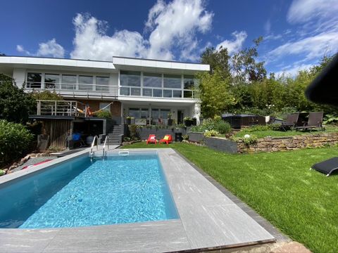 Ein modernes Haus mit großen Fenstern und einem gepflegten Garten. Im Vordergrund befindet sich ein Schwimmingpool, umgeben von Rasen und Liegestühlen.