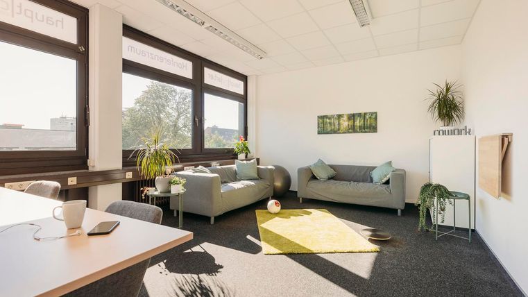 Ein helles Büro mit großen Fenstern und einer gemütlichen Sitzecke. Es gibt Pflanzen und einen Tisch mit einer Tasse und einem Notebook.