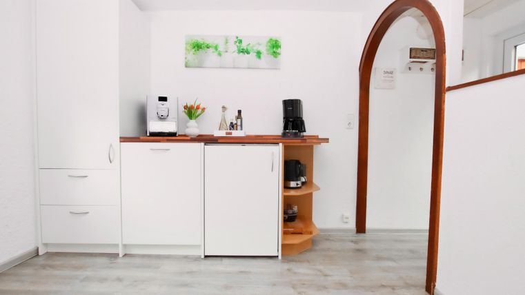Eine moderne Küchenzeile mit weißen Schränken und einem Holzregal. Auf der Arbeitsfläche stehen ein Kaffeegerät und eine Vase mit Blumen.