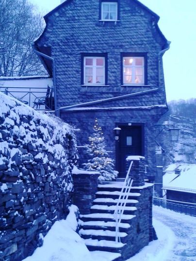 Ein charmantes Haus aus Stein in einer verschneiten Landschaft. Vor der Eingangstür steht ein kleiner Weihnachtsbaum.