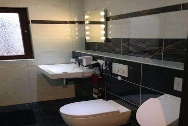 Ein modernes Badezimmer mit einer weißen Toilette und einem Schwimmbeckenwaschtisch. Die Wände sind hell gefliest, und ein großer Spiegel mit Beleuchtung ist vorhanden.