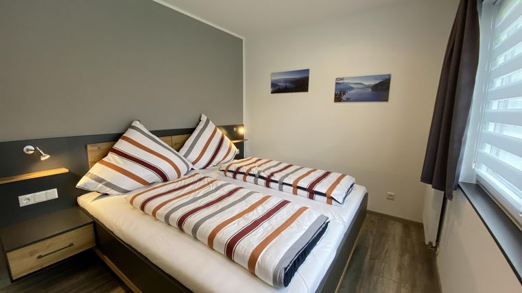 Ein modernes Schlafzimmer mit zwei Einzelbetten und gestreiften Bettdecken. Die Wände sind in neutralen Farben gehalten, und es gibt zwei Bilder sowie ein Fenster mit Gardinen.