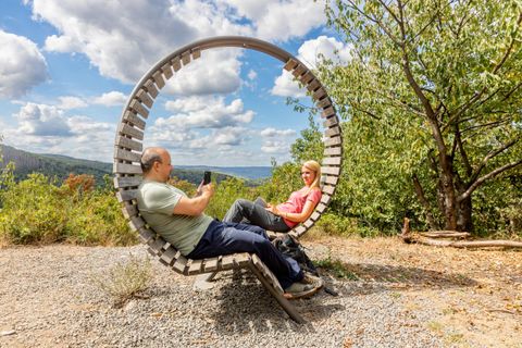 Ein Paar sitzt entspannt in einer runden Holzbank im Freien. Im Hintergrund sind Bäume und ein schöner Himmel mit Wolken zu sehen.