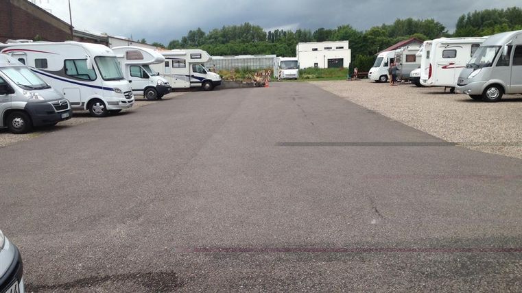Ein Parkplatz mit mehreren Wohnmobilen und einem grauen Asphaltboden. Im Hintergrund sind Bäume und ein Gebäude zu sehen, während der Himmel bewölkt ist.