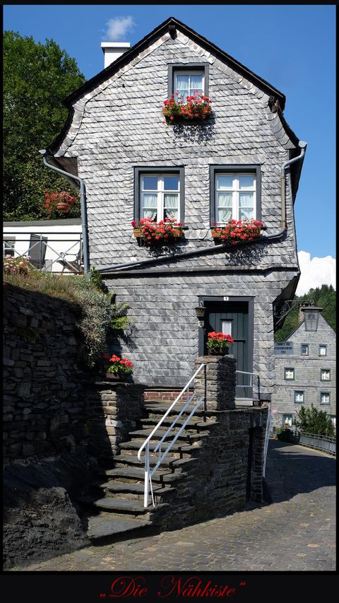 Ein charmantes Steinhaus mit blühenden Blumenkästen an den Fenstern. Die Umgebung ist ruhig und grün, ideal für einen gemütlichen Spaziergang.
