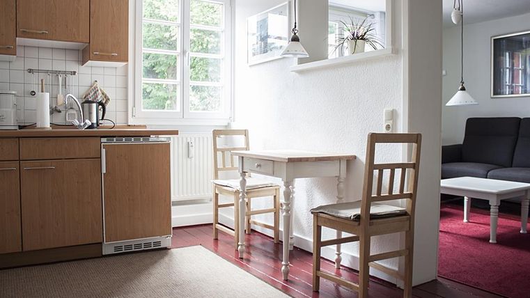 Eine gemütliche Küche mit Holzschränken und einem kleinen Essbereich. Ein leerer Tisch mit zwei Stühlen und ein Wohnbereich im Hintergrund.