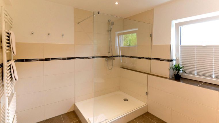 Ein modernes Badezimmer mit einer transparenten Dusche und hellen Fliesen. Naturalistische Dekorationen wie Pflanzen und Fenster sorgen für Helligkeit.