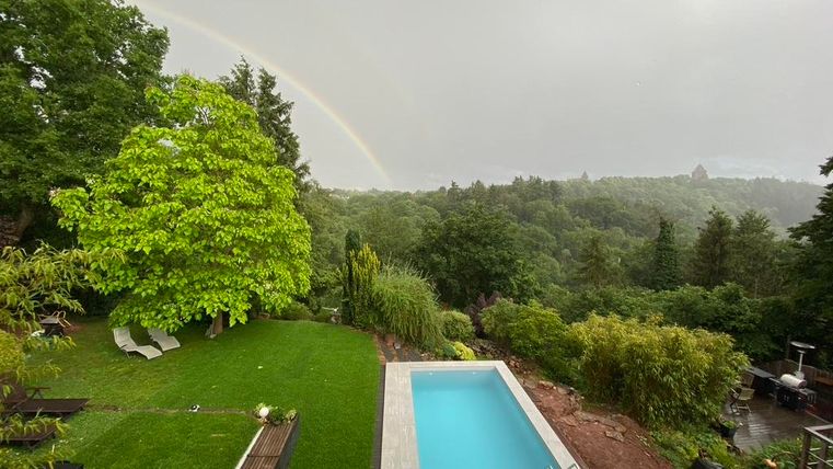 Ein großer Garten mit einem Swimmingpool und einem Regenbogen im Hintergrund. Die Umgebung ist grün und bewaldet.