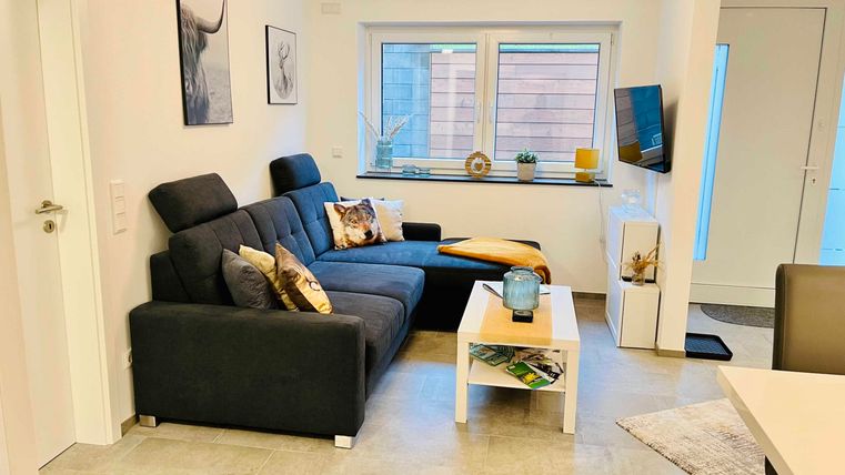 Ein modernes Wohnzimmer mit einer dunkelgrauen Couch und einem kleinen Tisch. Helle Wände und große Fenster sorgen für eine freundliche Atmosphäre.