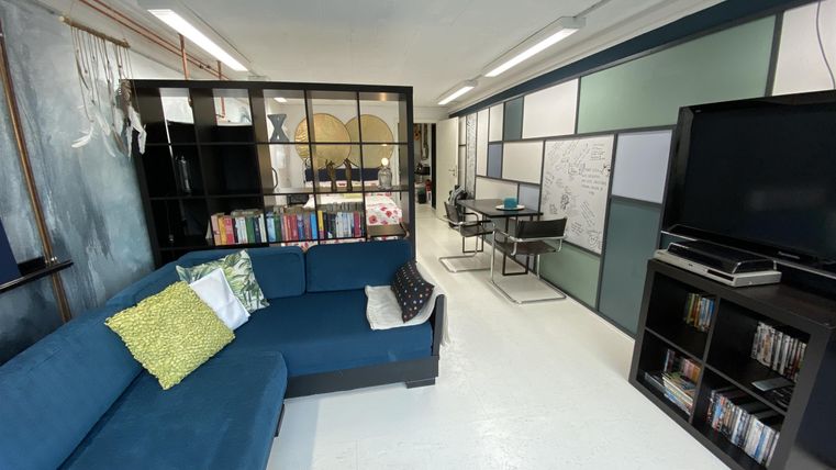 Ein modernes Wohnzimmer mit einem blauen Sofa und einem Bücherregal. Im Hintergrund sind Arbeitsplätze mit Computern und einer TV-Einheit zu sehen.