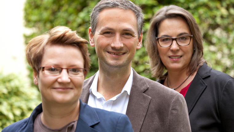 Drei Personen stehen lächelnd vor einer grünen Hintergrundkulisse. Sie tragen formelle Kleidung und wirken freundlich und professionell.