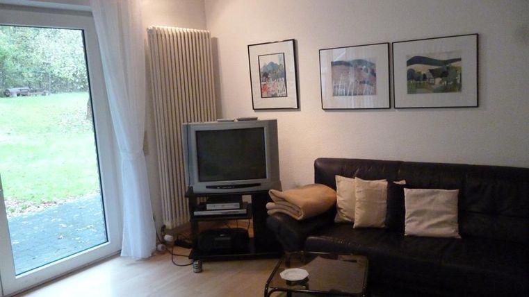 Ein gemütliches Wohnzimmer mit einem schwarzen Sofa und einem Fernseher. Große Fenster lassen viel Tageslicht herein und bieten einen Blick auf den Garten.