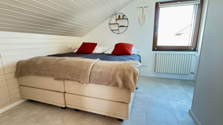 Ein gemütliches Schlafzimmer mit einem Doppelbett und zwei roten Kissen. Große Fenster sorgen für viel natürliches Licht.