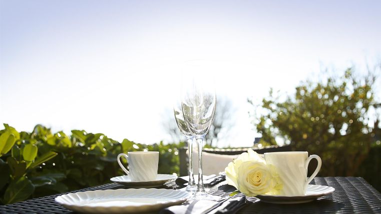 Ein elegant gedeckter Tisch im Freien mit weißen Tassen, Gläsern und einem Blumenstrauß. Der blauen Himmel und das grüne Blattwerk verleihen eine frische Atmosphäre.