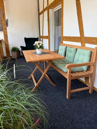 Eine gemütliche Sitzecke auf einem Balkon mit einem Holztisch und einer Bank. Auf dem Tisch steht eine kleine Blumenvase und im Hintergrund sind Pflanzen sichtbar.