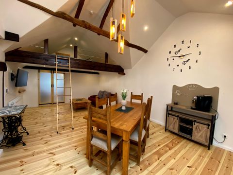 Ein helles Wohnzimmer mit Holzfußboden und freiliegenden Balken. Ein großer Esstisch und moderne Beleuchtung schaffen eine einladende Atmosphäre.
