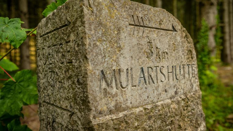 Ein steinerner Wegweiser mit der Inschrift "MULARTHÜTTE". Umgeben von Bäumen und pflanzlichem Grün.