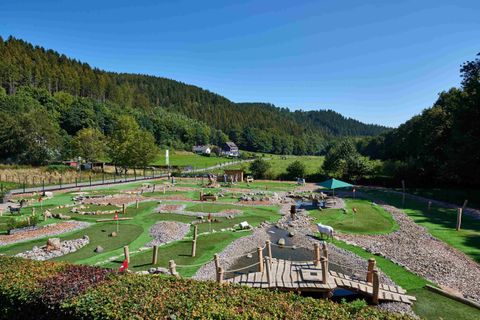 Ein weitläufiger Naturspielplatz in einer grünen Umgebung. Der Himmel ist klar und blau, umgeben von sanften Hügeln und Bäumen.