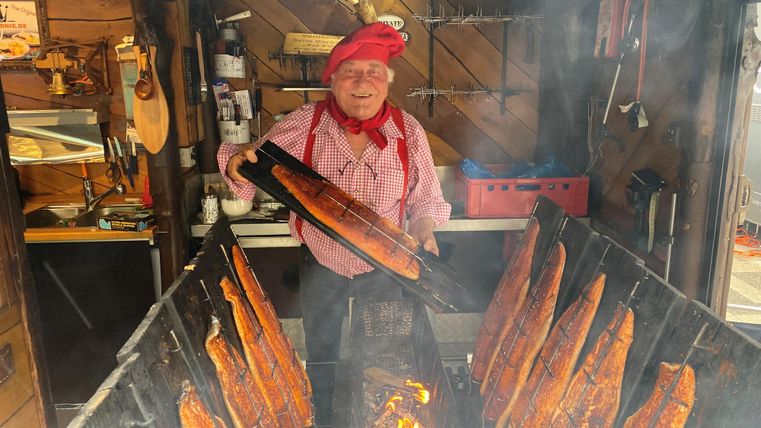 Ein Koch in traditioneller Kleidung steht vor einem Grill mit geräuchertem Fisch. Der Rauch verbreitet sich in der Luft und verleiht der Szene eine authentische Atmosphäre.