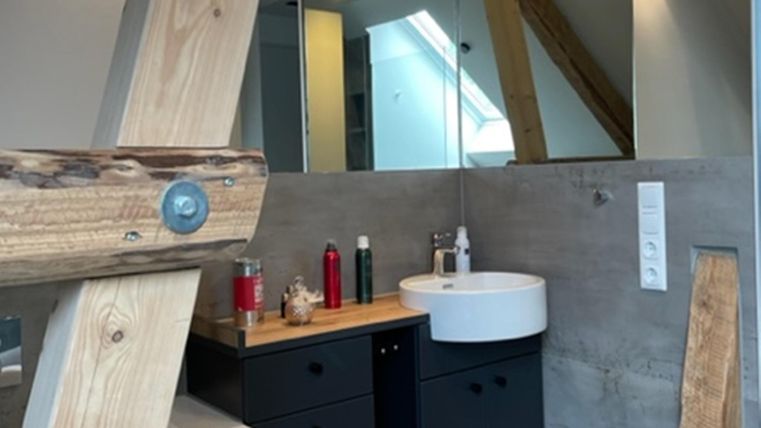 Ein modernes Badezimmer mit einer runden Waschbecken und Holzregal. Die Wände sind aus Beton, und es gibt große Fenster, die viel Licht reinlassen.