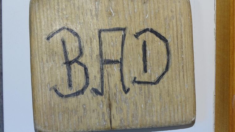 Ein Holzschild mit der Aufschrift "BAD". Es hängt an einer Wand und zeigt den Zugang zu einem Badezimmer an.