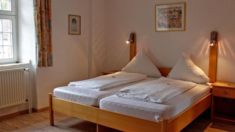 Ein helles Zimmer mit zwei Einzelbetten und Holzmöbeln. Die Wände sind neutral gestrichen und der Raum hat große Fenster, die für natürliches Licht sorgen.