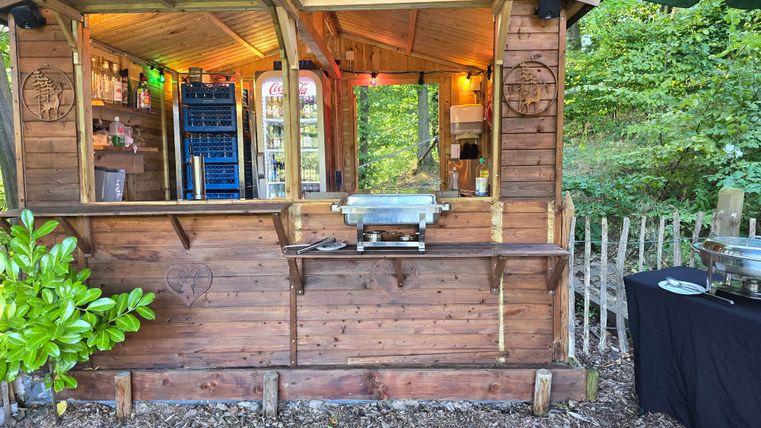 Ein Holzstand mit Fenster und Beleuchtung in einem grünen Umfeld. Auf dem Tisch steht ein Buffet mit verschiedenen Speisen.