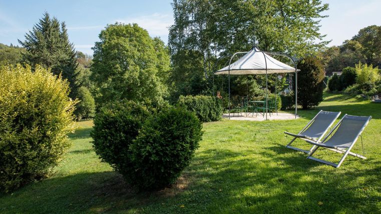 Eine grüne Wiese mit Sträuchern und einem Pavillon. Zwei Liegestühle laden zum Entspannen ein.