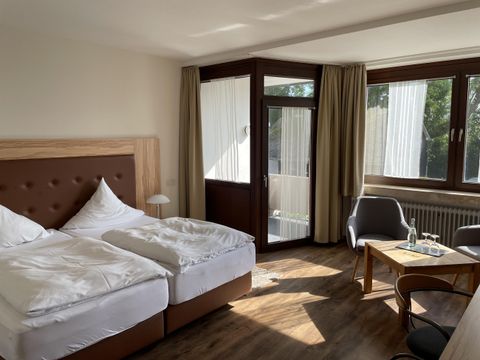 Ein helles Hotelzimmer mit zwei Einzelbetten, einem Tisch und Stühlen. Große Fenster sorgen für viel Tageslicht.