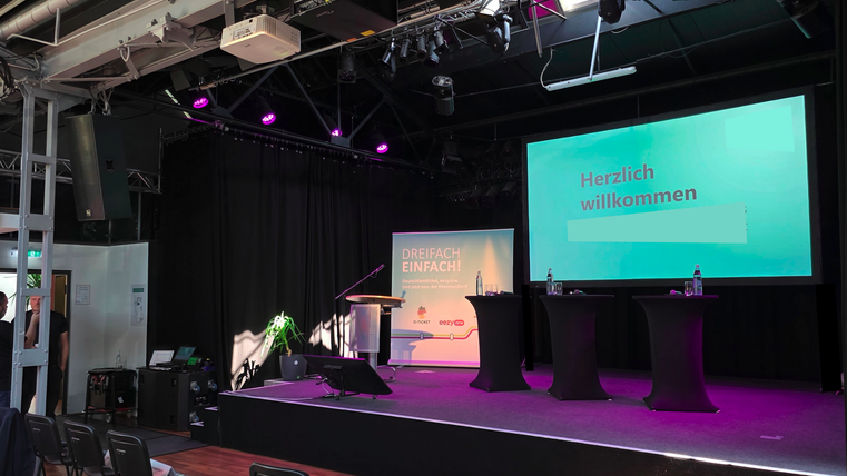 Eine Bühne mit einem Podium und zwei Stehtischen. Im Hintergrund befindet sich ein Bildschirm mit der Aufschrift "Herzlich willkommen".