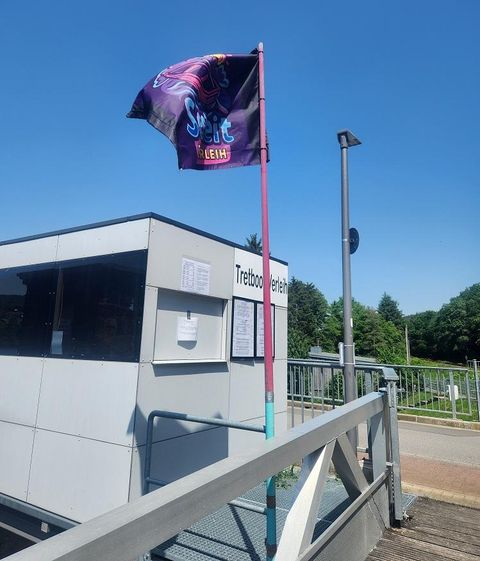 Ein Kiosk am Wasser mit einer bunten Fahne weht im Wind. Im Hintergrund sieht man Bäume und einen blauen Himmel.