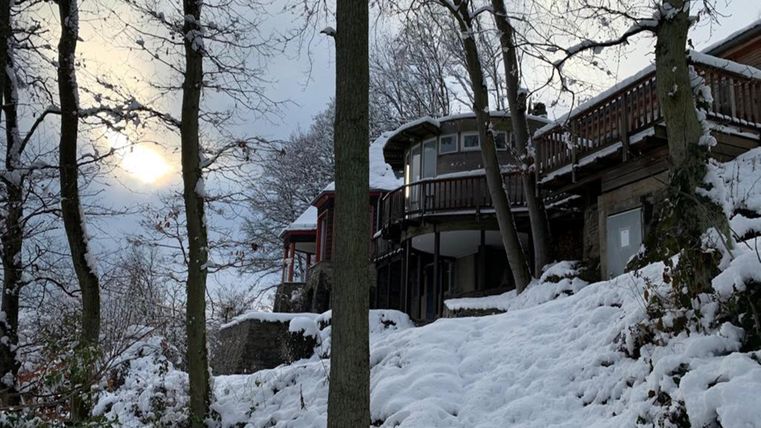 Eine verschneite Landschaft mit einem Haus, das auf einem Hügel steht. Die Sonne scheint schwach durch die Wolken.