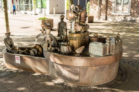 Eine kreative Brunnenanlage mit bronzeartigen Figuren und verschiedenen Motiven. Die Szene zeigt spielende Kinder und eine romantische Stadtlandschaft.