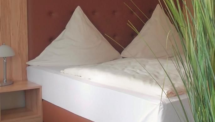 Ein gemütliches Doppelbett mit weißen Kopfkissen und einer hellen Bettdecke. Neben dem Bett steht ein Nachttisch mit einer Lampe und grünen Pflanzen im Hintergrund.